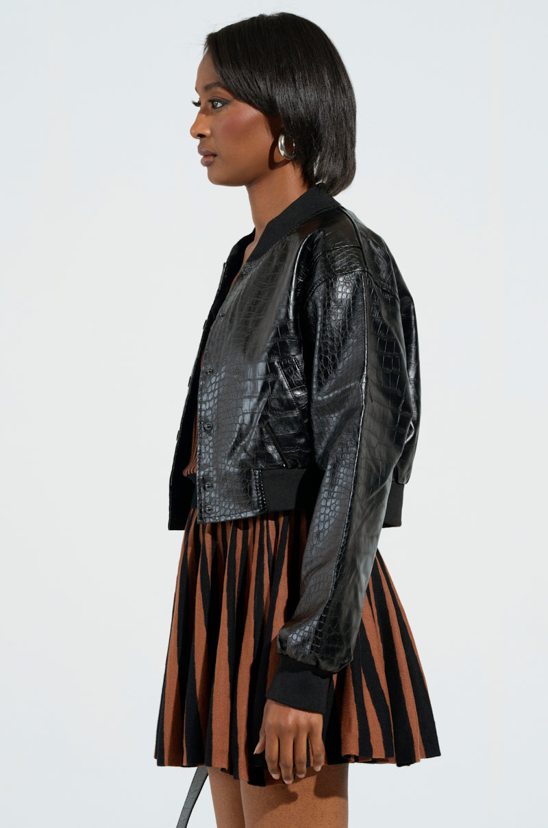 A person in profile poses against a plain background, wearing the WHAT I WANT FAUX LEATHER BOMBER, an embossed snakeskin-patterned black jacket, over a brown and black striped dress. Their hair is shoulder-length, and they have hoop earrings.