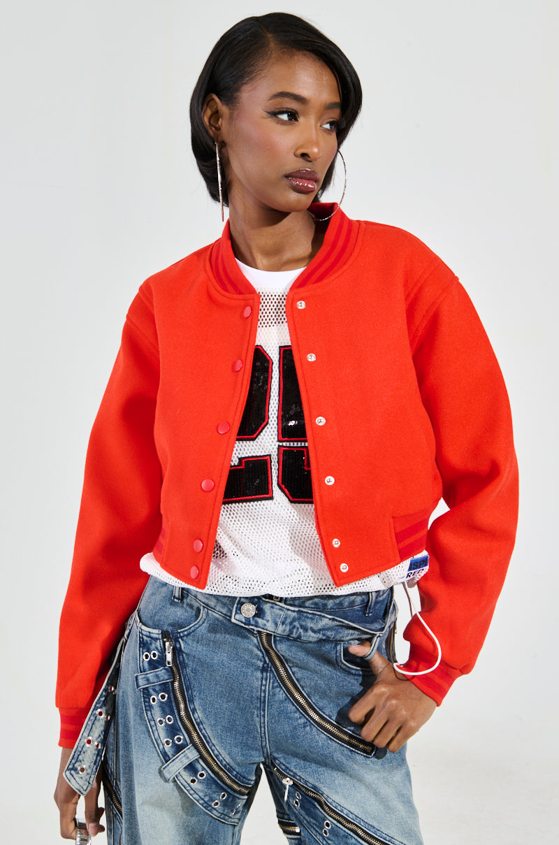 A person wearing the DAVIS EVERYDAY VARSITY BOMBER IN RED over a white mesh shirt featuring a black number. They accessorize with large hoop earrings and don jeans adorned with intricate strap details, giving off a sporty-chic vibe as they glance to the side against a plain background.