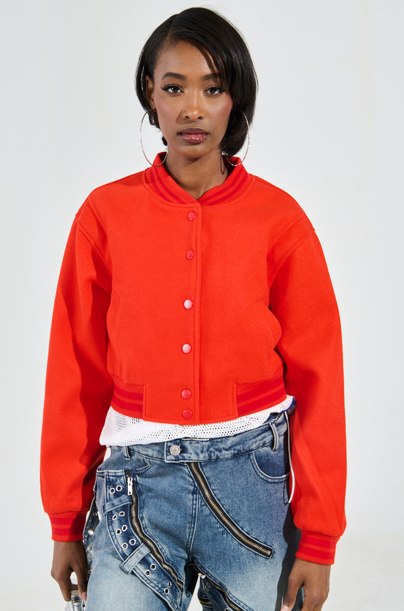 A person wearing the DAVIS EVERYDAY VARSITY BOMBER IN RED over a white shirt and blue jeans stands against a plain background. The jacket features buttons, while the jeans are accented with metal zipper details. The individual sports hoop earrings and short hair, creating a stylish blend of casual and chic.