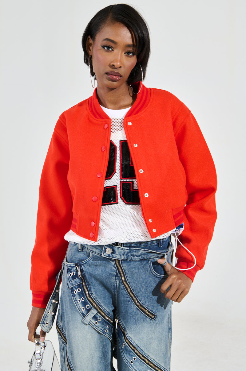 A person dressed in the DAVIS EVERYDAY VARSITY BOMBER IN RED over a white mesh top with a number, paired with zipper-accented jeans, stands against a plain background. They hold a transparent bag and wear hoop earrings, maintaining a neutral expression.