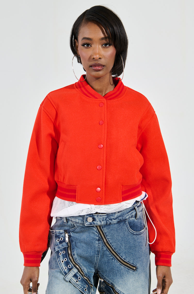 A person clad in the DAVIS EVERYDAY VARSITY BOMBER IN RED, paired with blue jeans, stands confidently against a plain background. They wear hoop earrings and exude an aura of self-assured style.