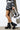 A person in a black and white plaid outfit holds a small handbag with the number 14. They wear AZALEA WANG DAXTON BLACK GLADIATOR FLATFORM SANDALS with a cutout design. The background is a white brick wall.