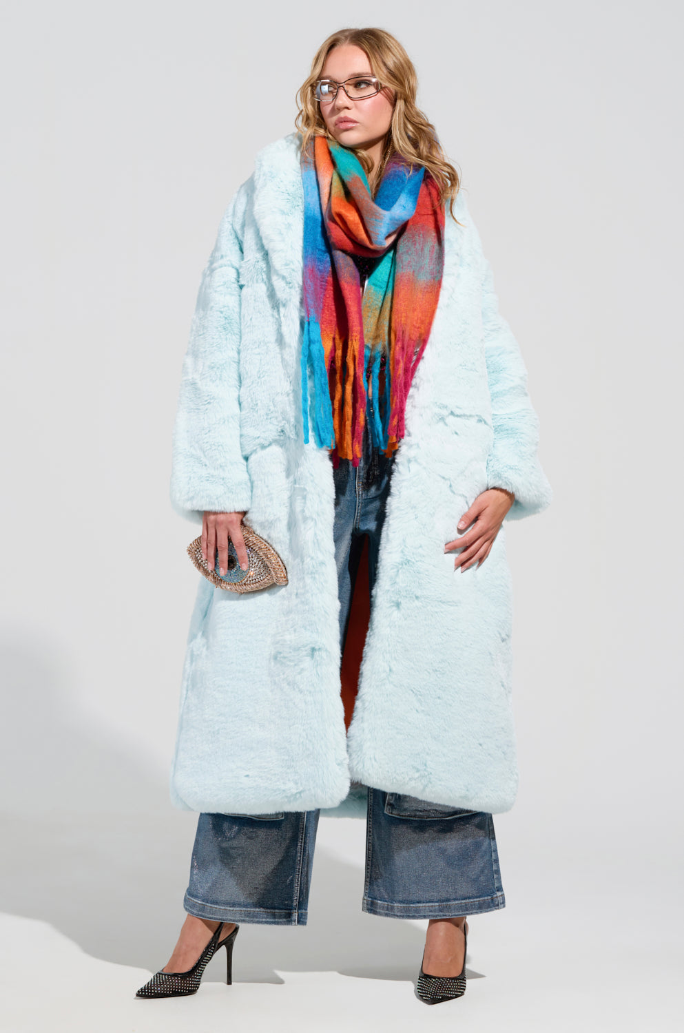 A person confidently stands wearing the DAYANNE Faux Fur Trench in light blue and a multicolored scarf. They have glasses, hold a small textured purse, and wear wide-leg jeans with black heels.