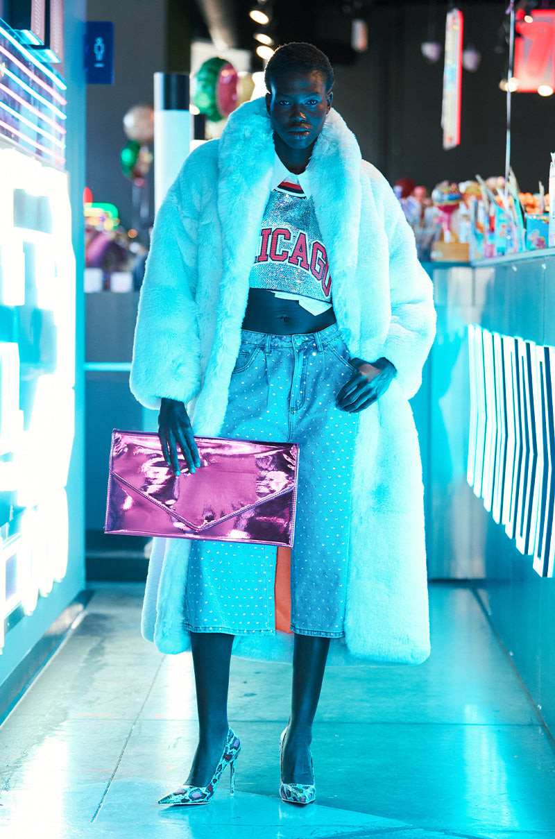 A person in the stylish Dayanne Faux Fur Trench in Light Blue stands confidently in a brightly lit space. They wear a Chicago crop top, denim skirt, and hold a shiny pink clutch. Neon lights and colorful decor create a vibrant atmosphere.