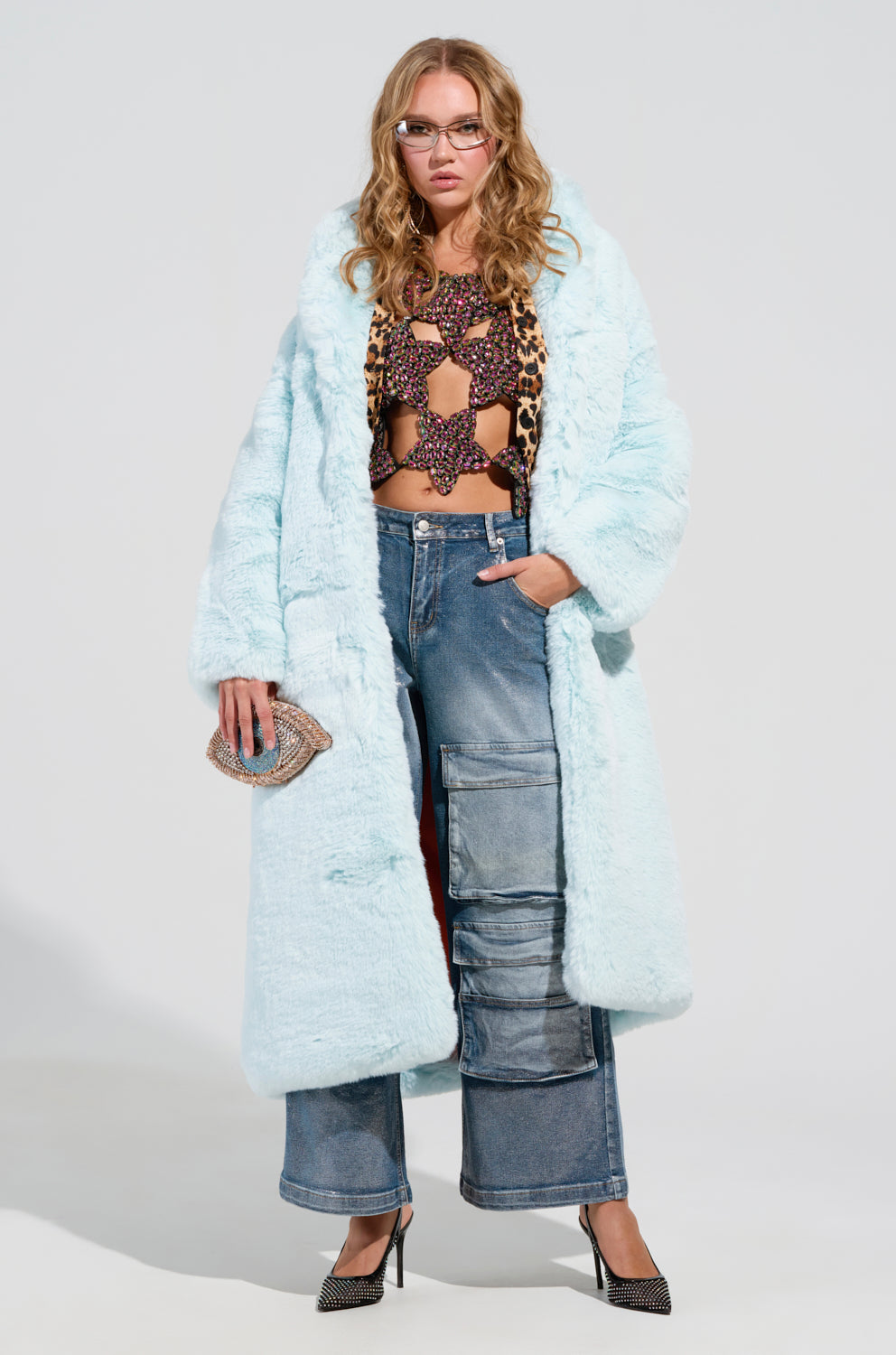 A person confidently stands wearing the DAYANNE FAUX FUR TRENCH IN LIGHT BLUE over a leopard print top. They're styled with wide-leg jeans and black high heels, holding a small sparkling clutch. Their look is completed with long wavy hair and glasses.