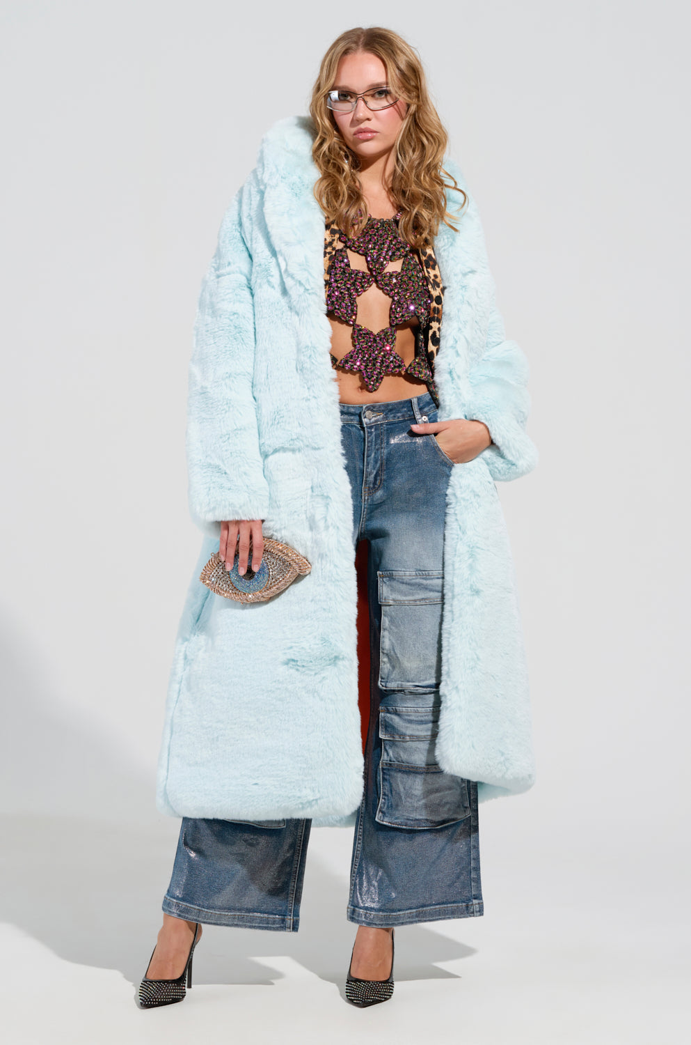 A woman wears the DAYANNE FAUX FUR TRENCH in light blue, layered over a colorful crochet top and leopard print pieces. Her wide-leg jeans enhance the oversized silhouette, as she accessorizes with stylish sunglasses, high heels, and a small purse against a plain background.