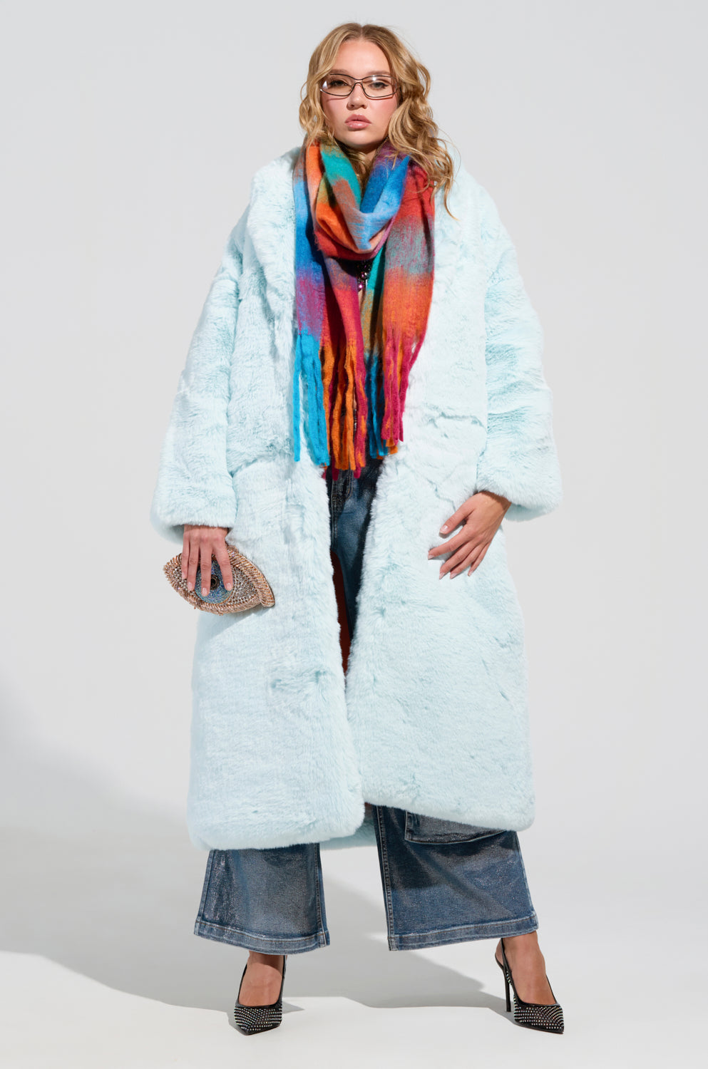 A person confidently stands wearing the DAYANNE Faux Fur Trench in Light Blue, paired with a colorful scarf and wide-leg jeans. They are holding a small, textured clutch and sporting black high-heeled shoes. The oversized silhouette beautifully contrasts against the plain white background.