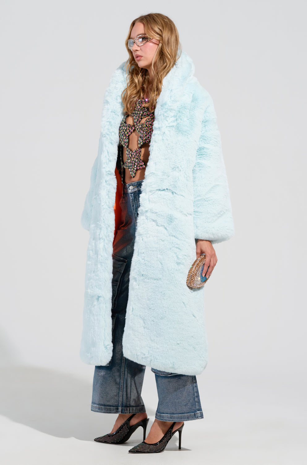 A person wrapped in the chic oversized silhouette of the Dayanne Faux Fur Trench in Light Blue drapes it over a colorful, patterned top and high-waisted jeans. They gracefully hold a small clutch bag while sporting glasses and high-heeled shoes, all set against a plain white background, which highlights the elegance of the trench.