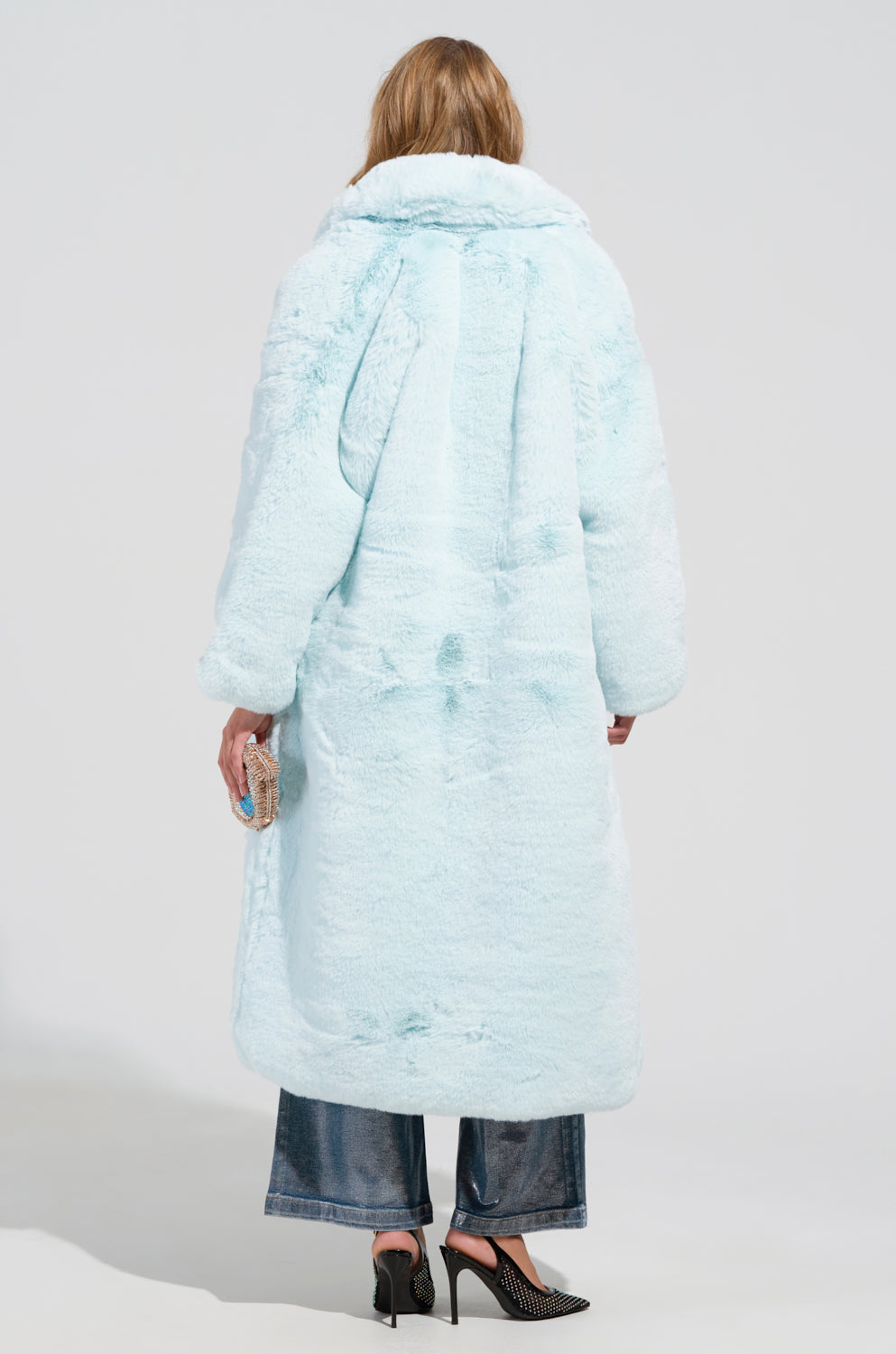 A person with blonde hair faces away, enveloped in the oversized silhouette of the DAYANNE FAUX FUR TRENCH IN LIGHT BLUE. They grasp a small, textured purse in their right hand and sport loose-fitting blue jeans paired with chic black high-heeled shoes against a simple, light gray backdrop.