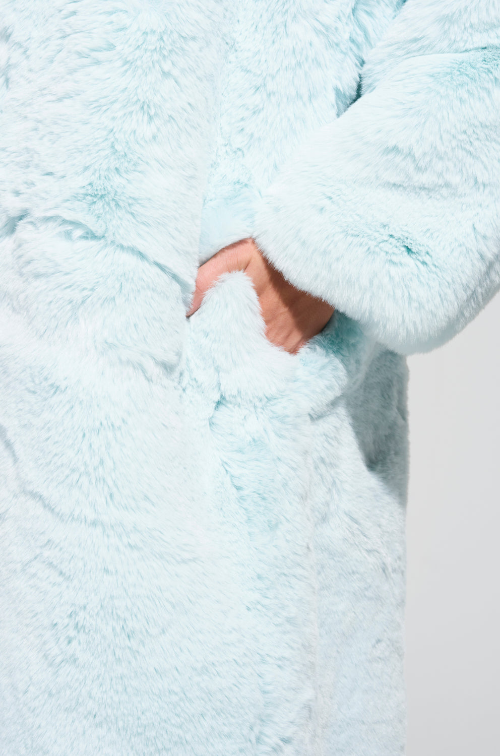 The Dayanne Faux Fur Trench in light blue features an oversized silhouette, with a person tucking their hand into one of the pockets. The close-up image emphasizes the texture of its soft, furry material.