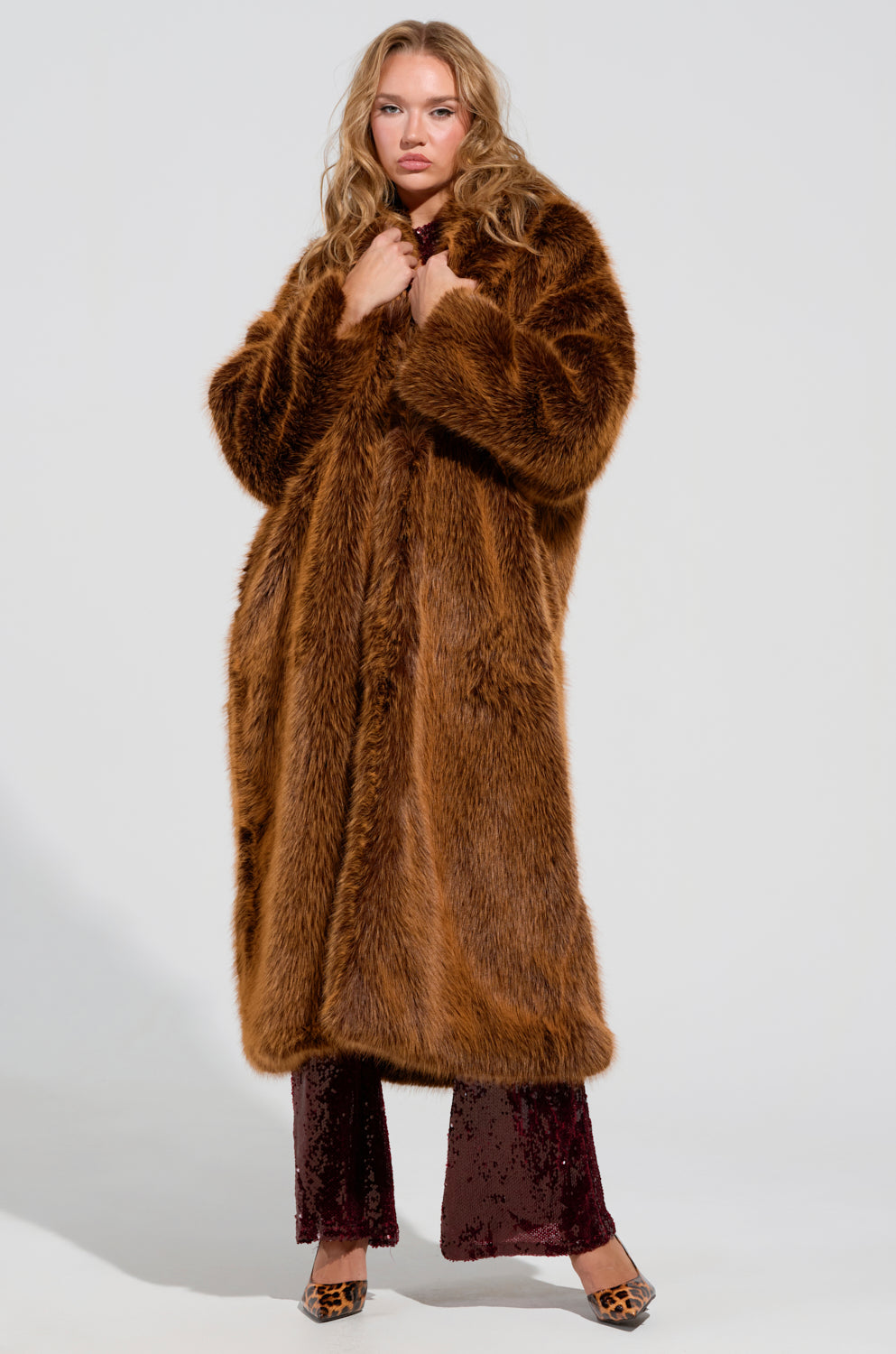 A person wearing the DAYANNE FAUX FUR TRENCH IN BROWN, which is maxi-length, stands against a simple backdrop. They are outfitted in dark pants and patterned shoes, seemingly clasping the coat closed at the neckline with both hands.