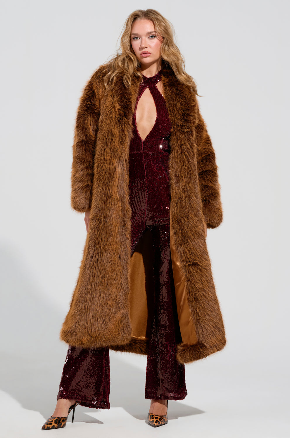 A person stands confidently with hands on hips, wearing the DAYANNE FAUX FUR TRENCH in brown over a burgundy sequin jumpsuit featuring a keyhole neckline and flared legs. The ensemble is completed with leopard-print heels, creating an oversized silhouette in their bold look.