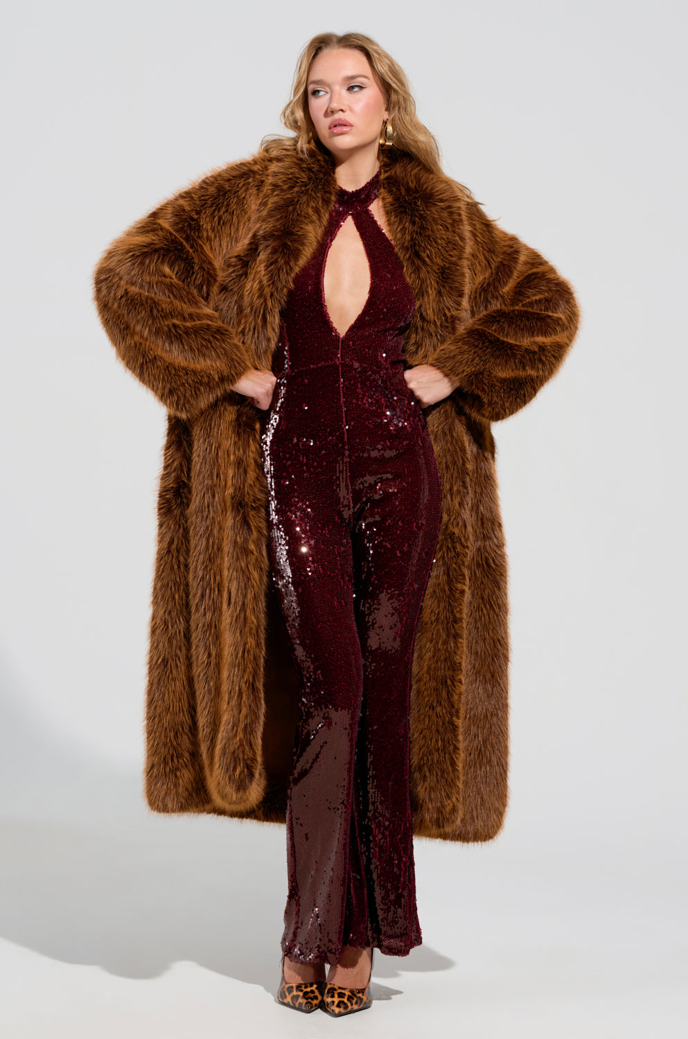 A woman poses confidently, draped in the DAYANNE Faux Fur Trench in brown over a sparkly, burgundy jumpsuit with a halter neckline. She stands against a plain white background, her left hand on her hip. Completing the look are her leopard print heels.