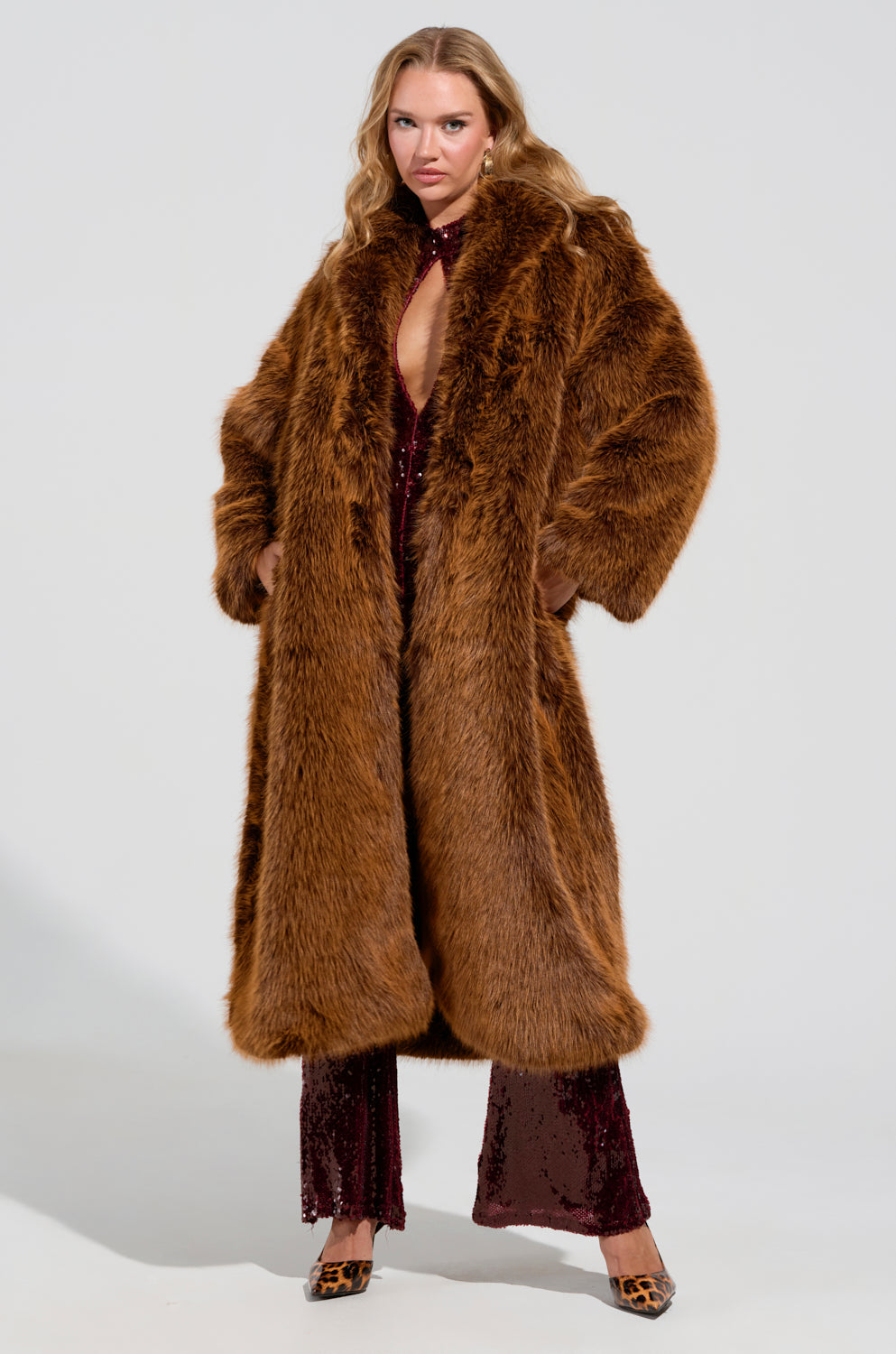 A woman poses confidently in a DAYANNE Faux Fur Trench in brown over a sparkling burgundy outfit and leopard print shoes. She has long, wavy blonde hair and is standing against a plain white background.