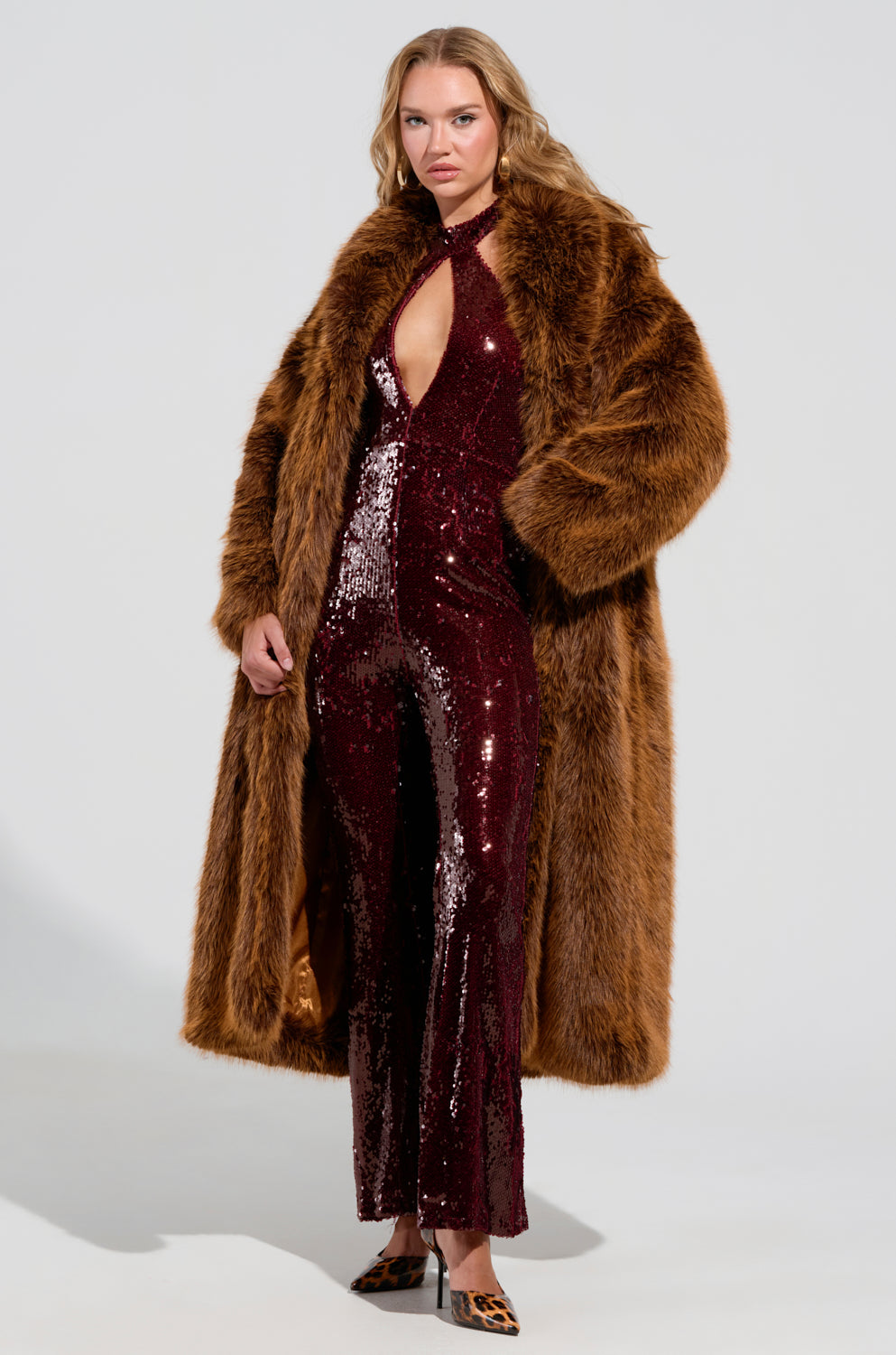 Draped in the DAYANNE FAUX FUR TRENCH IN BROWN, a person stands confidently donning a sequined burgundy jumpsuit with a keyhole neckline, their hands relaxed by their side as they showcase leopard-patterned shoes. The oversized silhouette adds drama against the plain white background.