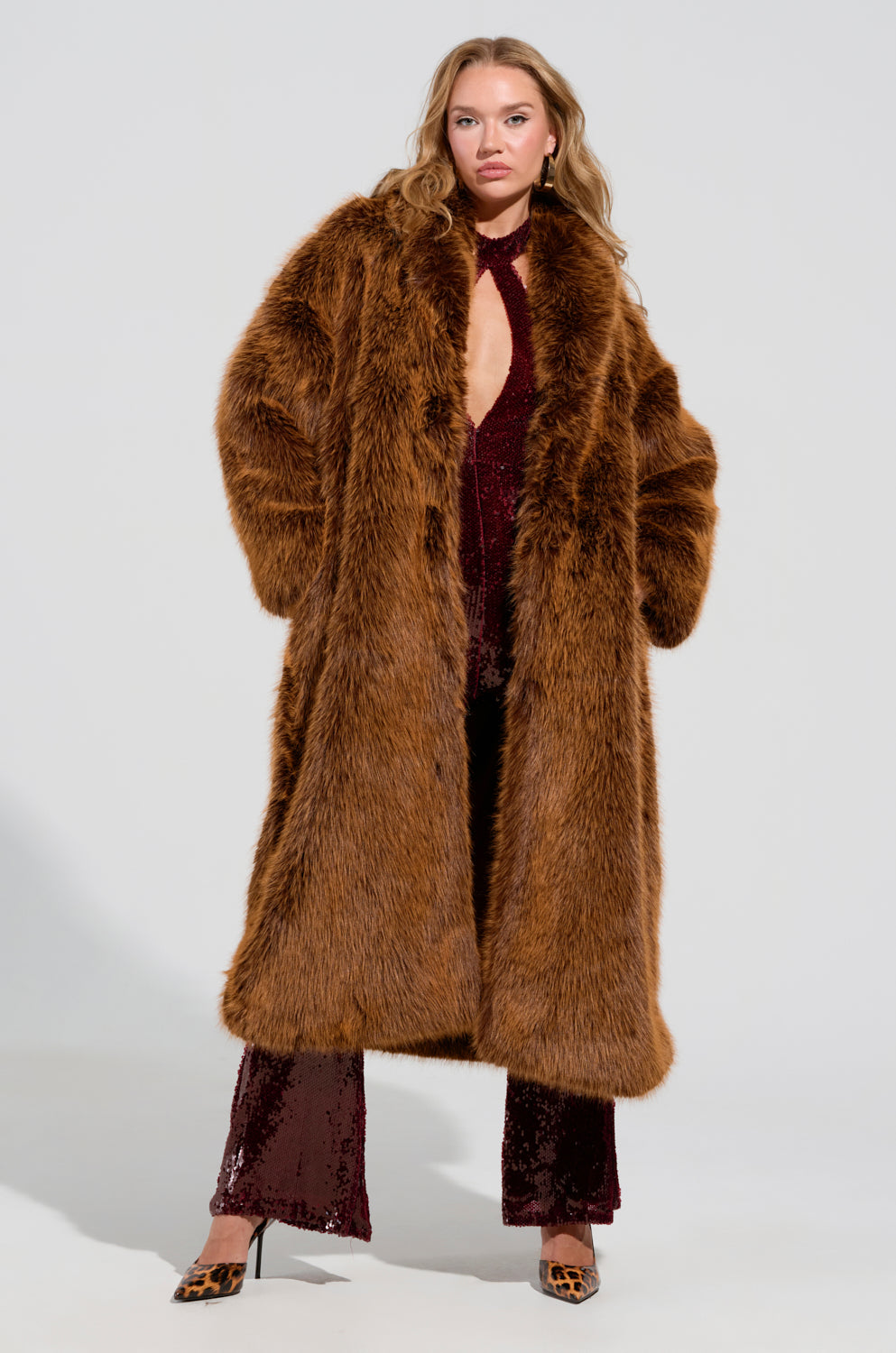 A person wearing the Dayanne Faux Fur Trench in brown stands confidently. Beneath the oversized silhouette, a sequined dark red jumpsuit and leopard-print heels are partially visible. The plain and light background highlights the outfit's rich textures and bold colors.