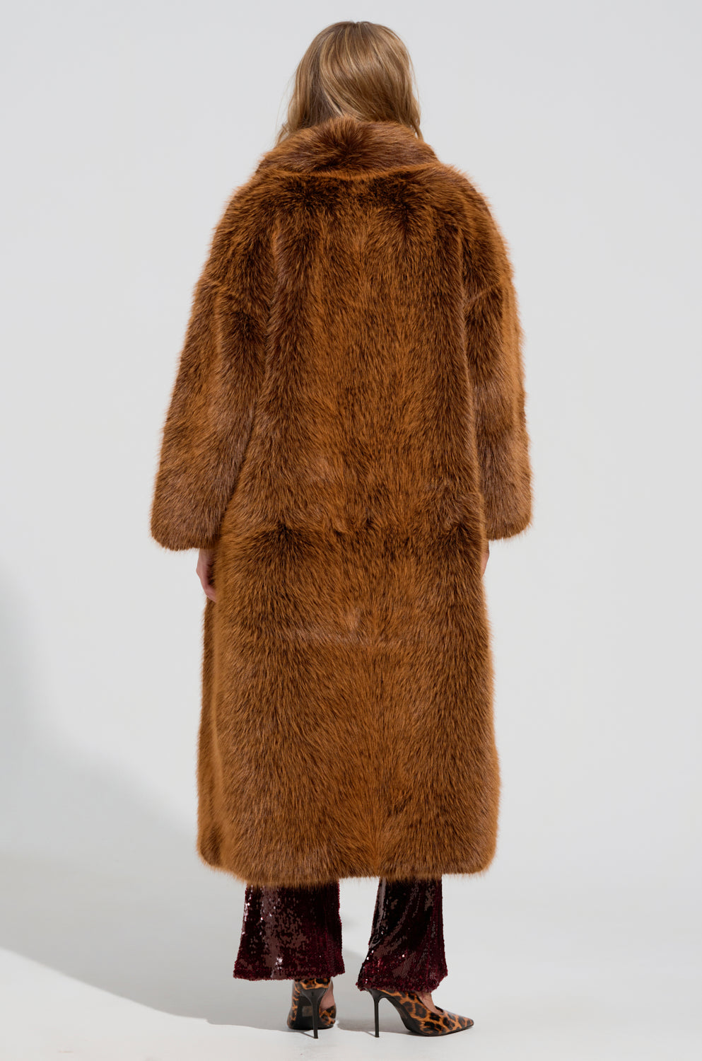 A person with long hair is seen from behind, wearing the luxurious DAYANNE Faux Fur Trench in brown, which falls gracefully to maxi-length. They pair it perfectly with dark pants and chic leopard print high heels. The plain, light-colored background provides a striking contrast to the oversized silhouette of this stylish ensemble.
