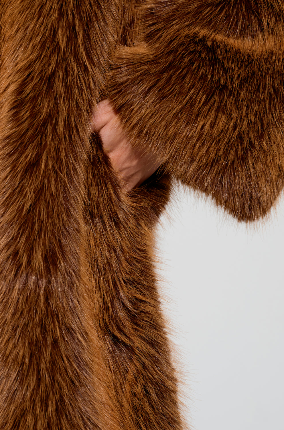 In the image, a person is seen wearing the DAYANNE FAUX FUR TRENCH IN BROWN. Their left hand is tucked into one of the coat's pockets, highlighting its rich color and luxurious texture that offers a warm appearance. The oversized silhouette enhances its chic allure against a plain, light-colored background.