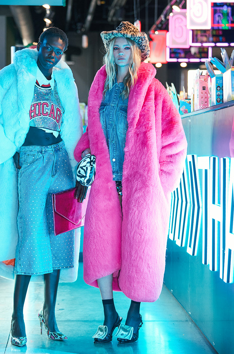 In a vibrant, neon-lit setting, two models exude confidence. One flaunts an AKIRA Label light blue faux fur trench over a Chicago top and jeans. The other dazzles in the DAYANNE Faux Fur Trench in Fuchsia over a denim outfit and cow-print hat as they stand near a brightly lit counter. 1