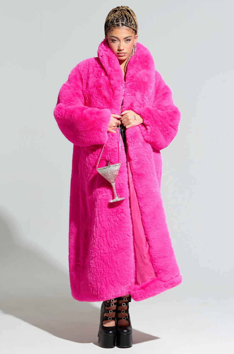 A person confidently wearing the DAYANNE FAUX FUR TRENCH in vibrant fuchsia stands out with a chic flair. They carry a sparkly, oversized goblet-shaped bag and sport platform boots with prominent laces against a plain, soft background. 3