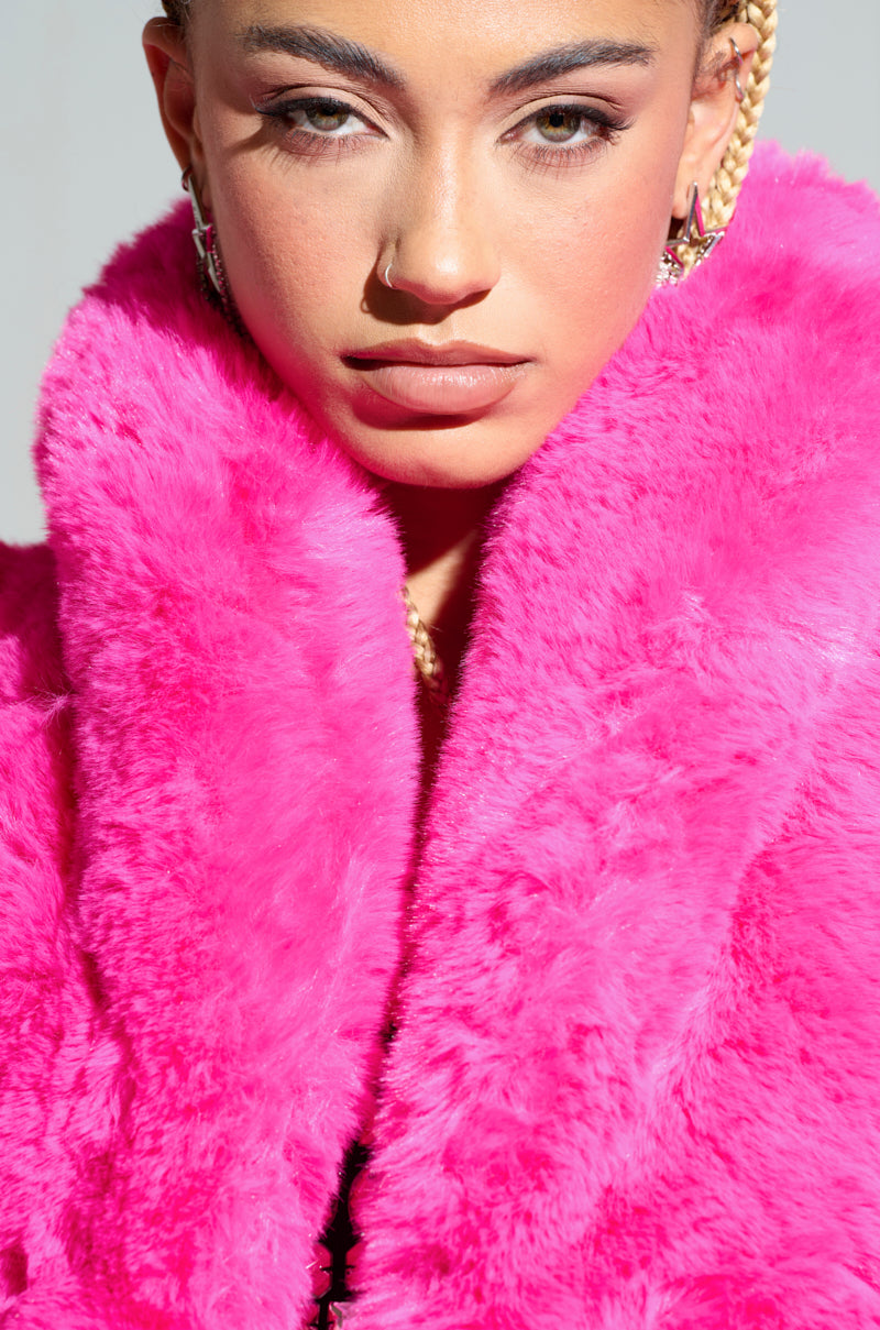 The individual is adorned in the DAYANNE FAUX FUR TRENCH IN FUCHSIA and gold hoop earrings, with their braided hair elegantly styled in an updo. Their serious expression contrasts against the neutral background, highlighting the vibrant hue of their chic outfit. 6