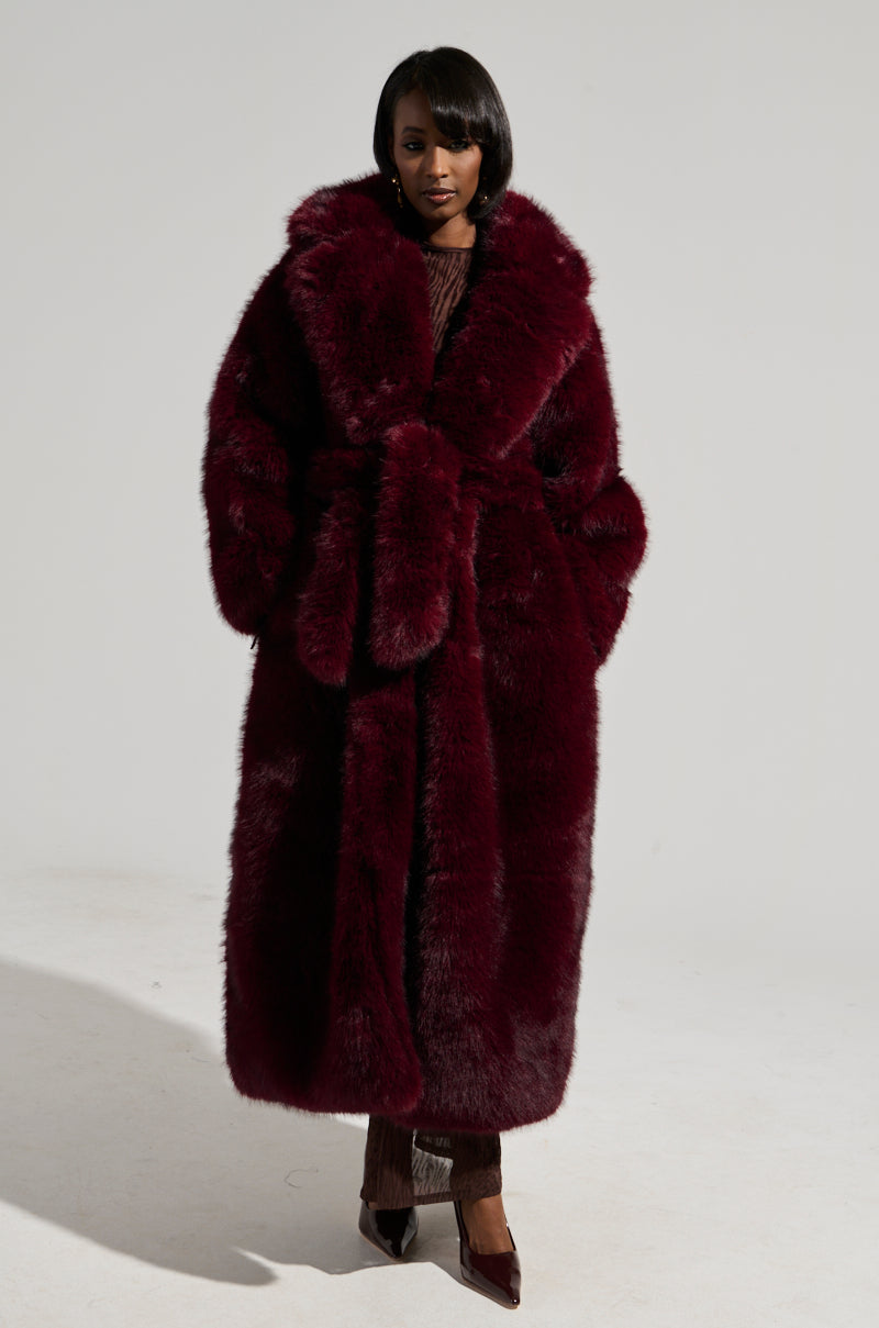 A person stands confidently in the DAYANNE FAUX FUR TRENCH IN WINE, a maxi-length coat with a matching belt. Their dark hair is styled neatly, and they wear heeled shoes. The deep wine color of the coat contrasts strikingly against the plain white background.