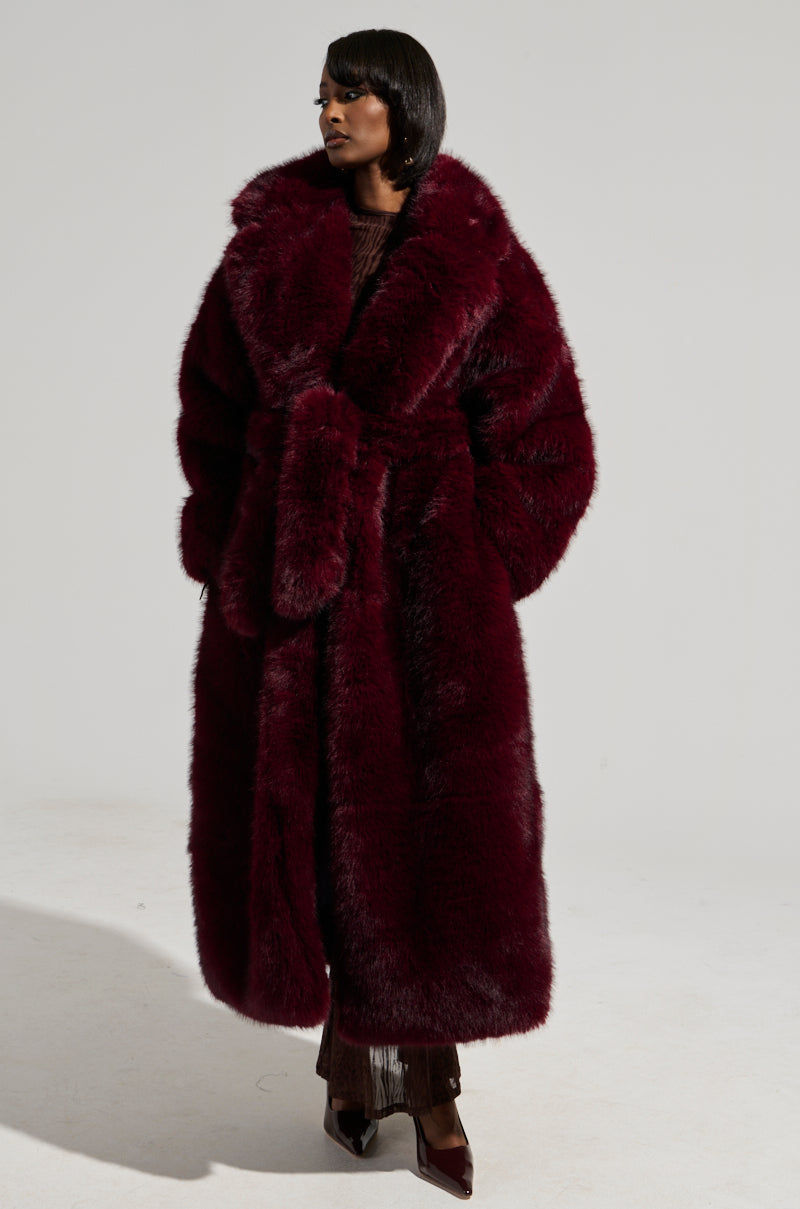 Clad in the DAYANNE FAUX FUR TRENCH IN WINE, an individual stands confidently against a plain background. With short, styled hair and pointed dark shoes, they exude elegance in every detail.