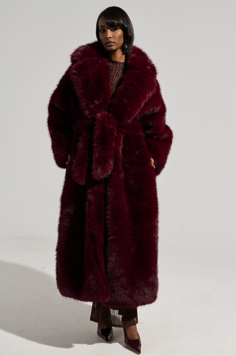 A person stands confidently, wearing the DAYANNE FAUX FUR TRENCH IN WINE with a matching belt. They are dressed in dark pointed shoes and pose against a plain background.
