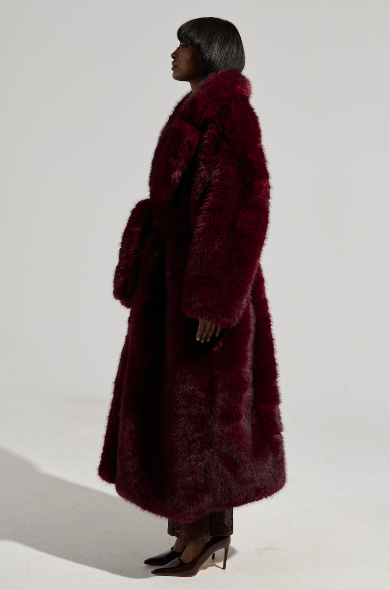 A person stands in profile, draped in the Dayanne Faux Fur Trench in Wine. Their hair is straight and dark, paired with sleek, pointed shoes. The plain off-white background serves to highlight the maxi-length coat’s commanding presence.
