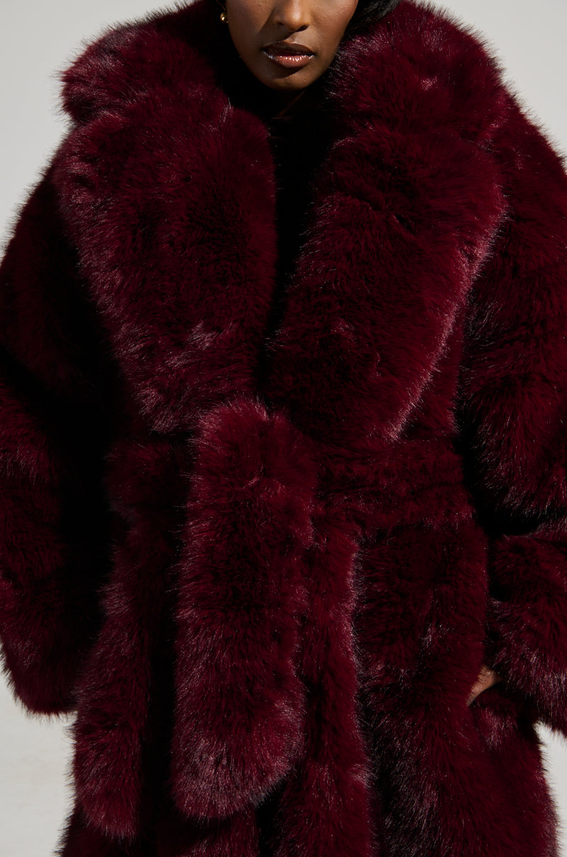 A person wearing the DAYANNE FAUX FUR TRENCH IN WINE, a luxurious maxi-length coat characterized by its rich wine-colored hue, large fluffy lapels, and matching belt. The soft, plush texture of the faux fur adds elegant volume to the outfit against a neutral background.