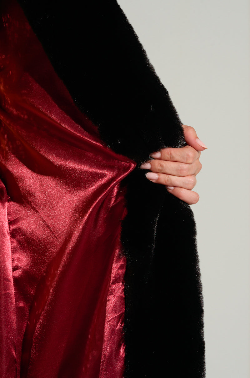 A hand holds open the DAYANNE LONG SATIN LINING FAUX FUR TRENCH COAT, showcasing its lustrous red satin lining. The focus is on the contrast between the dark faux fur exterior and the vibrant interior, enhanced by the longline silhouette that adds elegance to this striking garment.