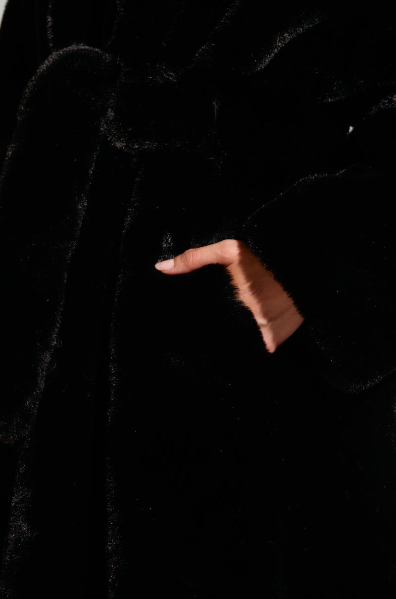 A close-up of someone wearing the DAYANNE LONG SATIN LINING FAUX FUR TRENCH COAT in black, showcasing its tied waist and longline silhouette. Their hand rests in a side pocket, highlighting the coat's soft and glossy texture, complemented by its luxurious satin lining.