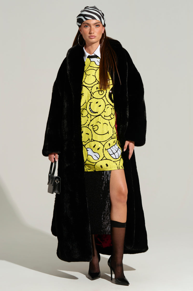 Dressed confidently in the DAYANNE LONG SATIN LINING FAUX FUR TRENCH COAT, its sleek satin lining subtly visible, a person wears a vibrant yellow dress adorned with smiley faces. A zebra-patterned headscarf adds flair, while hoop earrings glimmer brilliantly. They hold a stylish black handbag and complete their ensemble with knee-high socks and heels, all set against a plain backdrop.