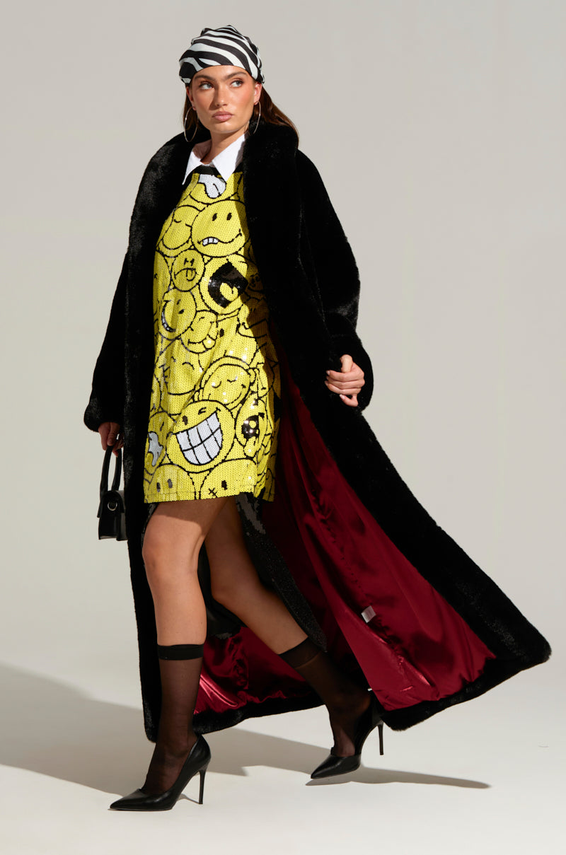 A fashionable person walks confidently wearing the DAYANNE Long Satin Lining Faux Fur Trench Coat in black, featuring a luxurious red satin lining, over a yellow dress adorned with smiley faces. They accessorize with a black and white striped headscarf, a small black bag, and sleek black heels.