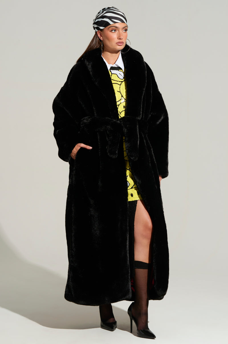 A person wears the DAYANNE LONG SATIN LINING FAUX FUR TRENCH COAT over a yellow patterned dress. They accessorize with a zebra-patterned headscarf and black heeled boots, posing gracefully against a plain background, with the coat's satin lining subtly peeking through.