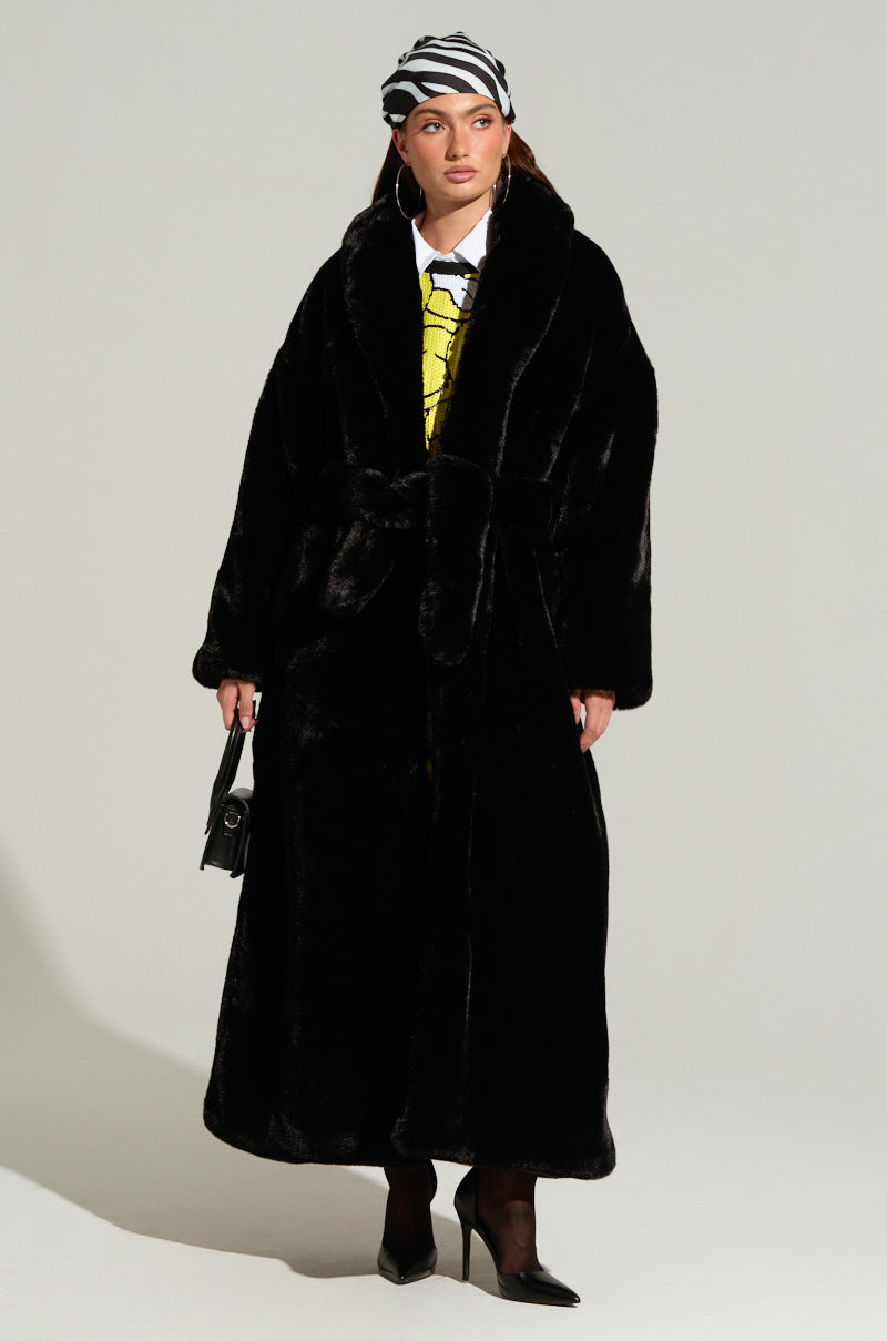 A person stands wearing the luxurious DAYANNE LONG SATIN LINING FAUX FUR TRENCH COAT in black, paired with a yellow shirt and black heeled shoes. They hold a small black handbag and don a black-and-white striped headscarf. The background is plain gray.