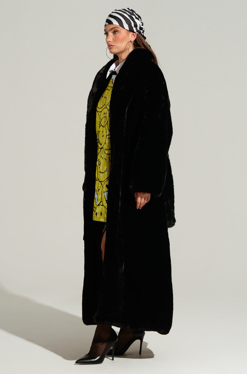 Against a simple backdrop, an individual showcases an elegant ensemble with a DAYANNE LONG SATIN LINING FAUX FUR TRENCH COAT in a luxurious black finish, layered over a yellow patterned dress. The look is accentuated by a zebra-striped headscarf and chic black high heels, exuding sophistication in every element.