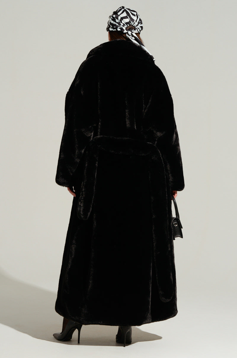 Clad in the DAYANNE Long Satin Lining Faux Fur Trench Coat, a person stands facing away, paired with black high-heeled boots and a black and white patterned headscarf, while holding a small black bag against a minimalist background.
