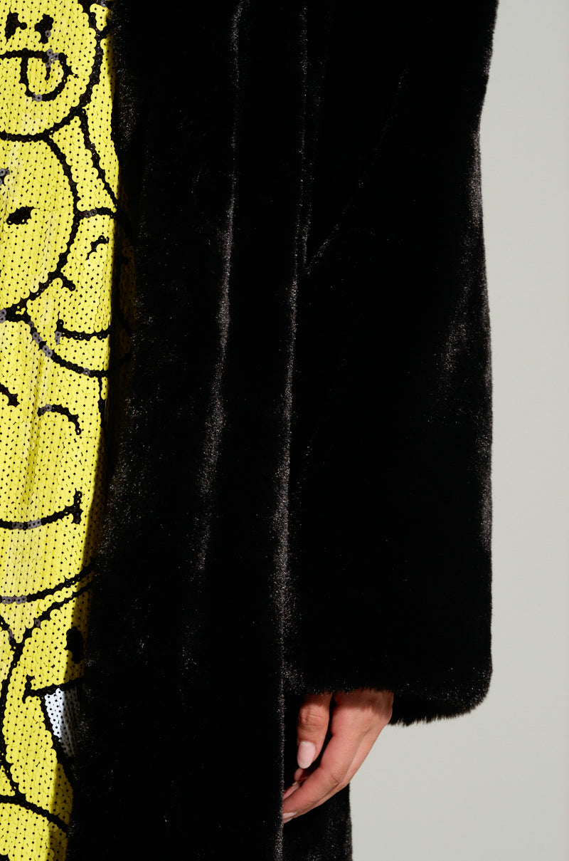 A person wearing the DAYANNE LONG SATIN LINING FAUX FUR TRENCH COAT in black, showcasing its longline silhouette over a yellow outfit with black smiley face patterns. The partially visible hand draws attention to the coat's soft texture and satin lining against a plain, light background.