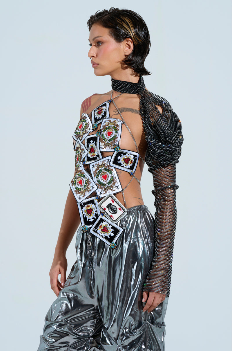 A person stands against a plain background, dressed in a standout AKIRA Label ensemble that features the ADDED DRAMA RHINESTONE SLEEVE along with playing cards and metallic, reflective pants. They sport short, slicked-back hair.