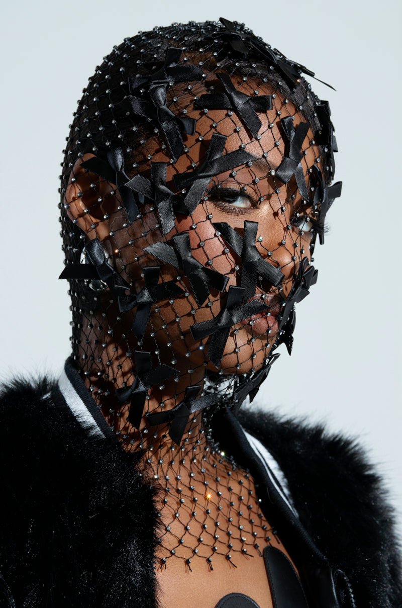 A person is wearing the "IM JUST A GURL BOW BALACLAVA," a decorative netted headpiece embellished with rhinestones. The ensemble features a black fur garment that contrasts with the balaclava's elaborate dark fishnet design covering the face and head.