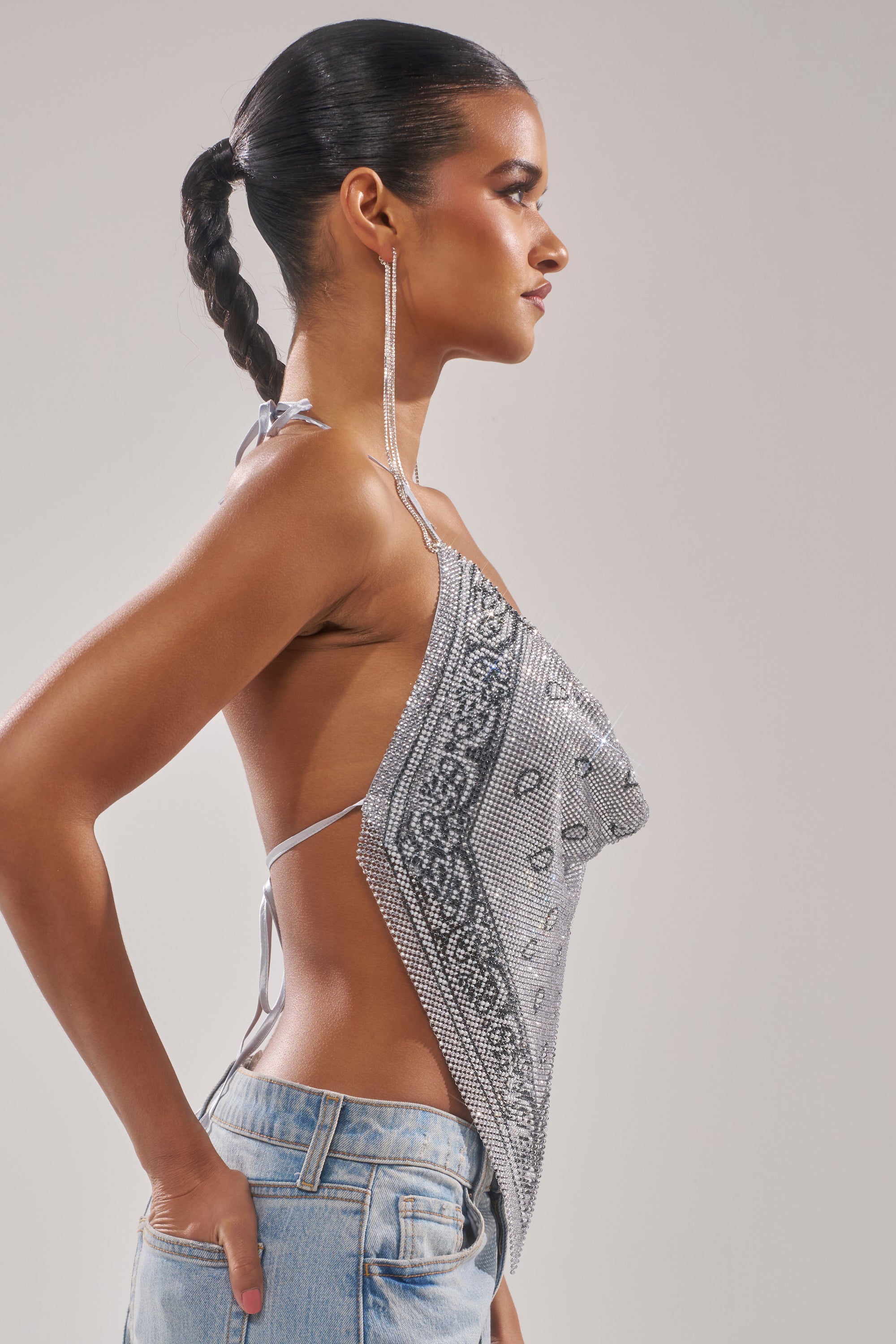 A woman with a sleek braided ponytail models the FREESTYLE RHINESTONE COWL NECK TOP and light blue jeans, posing in profile with hands on hips against a plain light background. 3