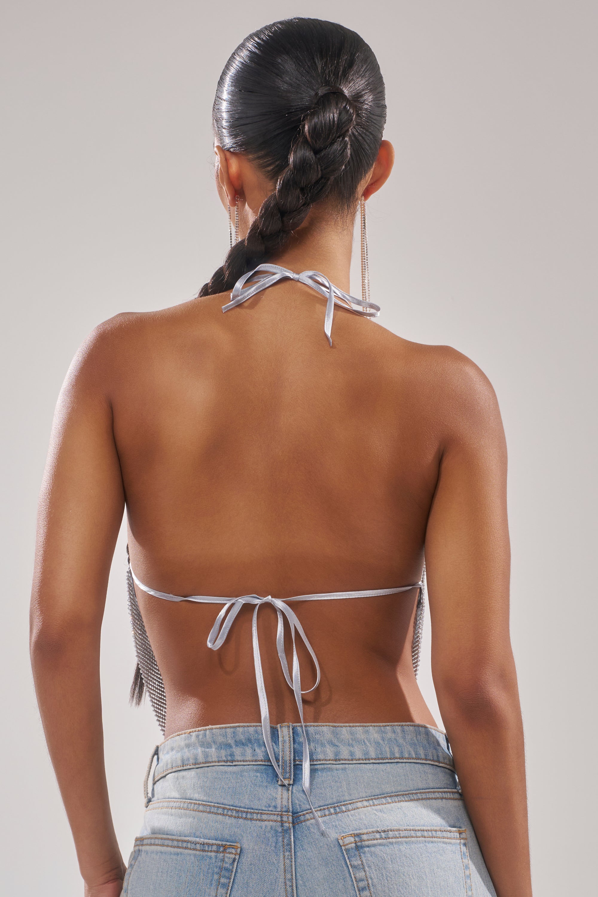 A woman with a long, braided ponytail is shown from the back wearing the FREESTYLE RHINESTONE COWL NECK TOP, which features a halter-style tie-back and leaves her back mostly exposed. She pairs it with light blue jeans against a plain background. 4