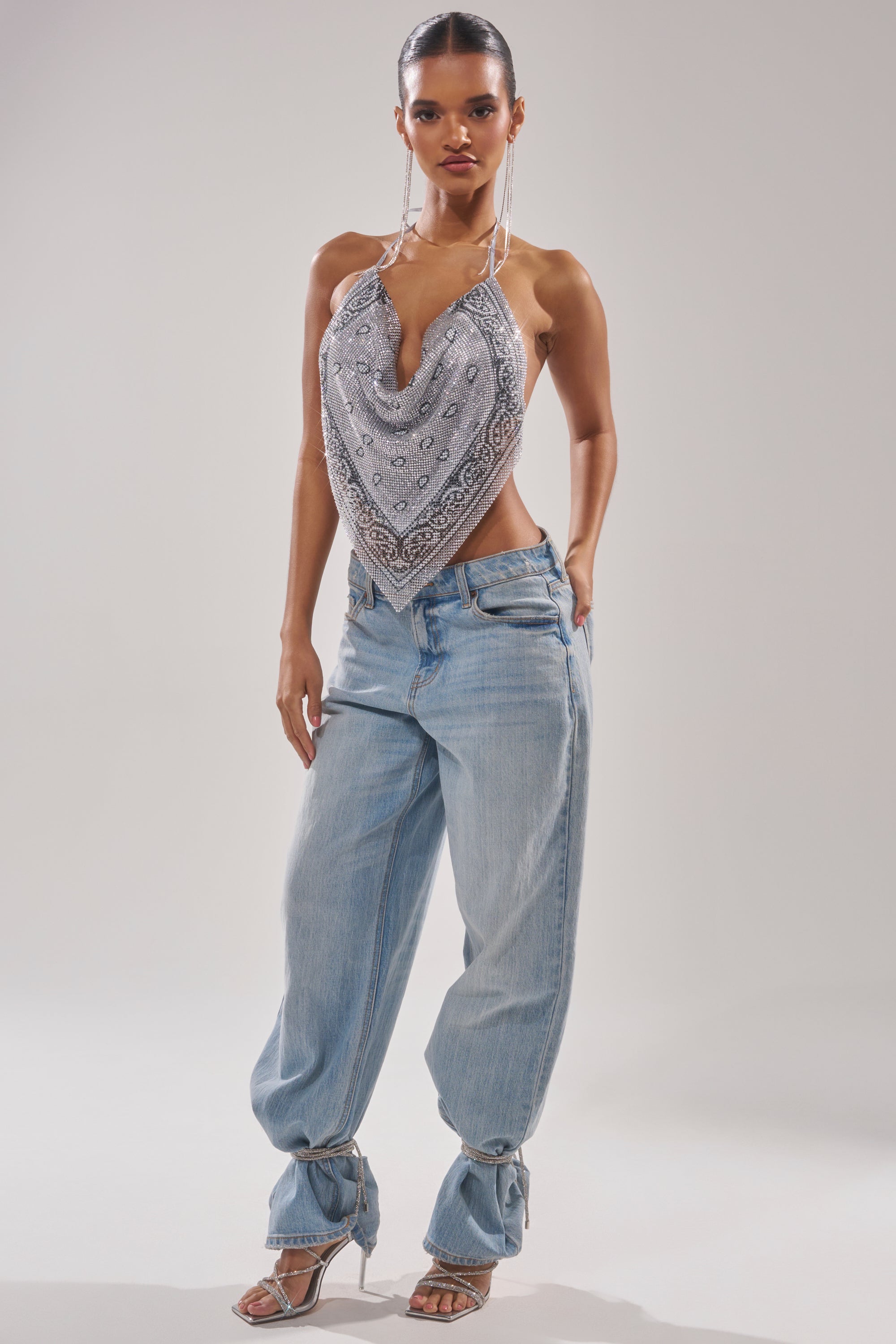 A woman poses in the FREESTYLE RHINESTONE COWL NECK TOP, light-wash loose jeans with tied ankles, hoop earrings, and strappy heels—an effortlessly cool concert festival outfit against a plain background. 6