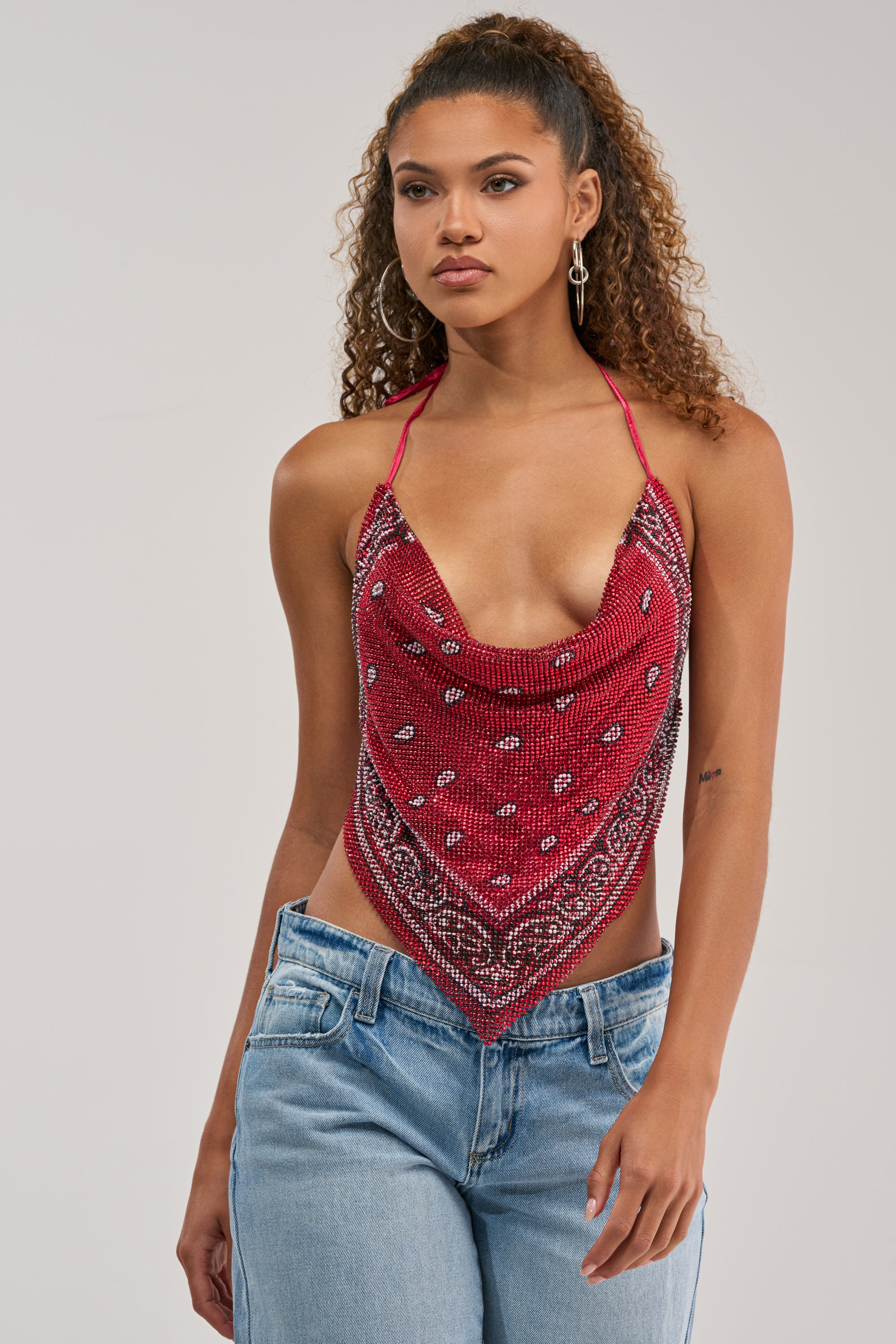A woman with curly hair and large hoop earrings wears the FOREVER AND EVER COWL NECK BLOUSE in a red paisley pattern with light blue jeans, standing against a plain light background.