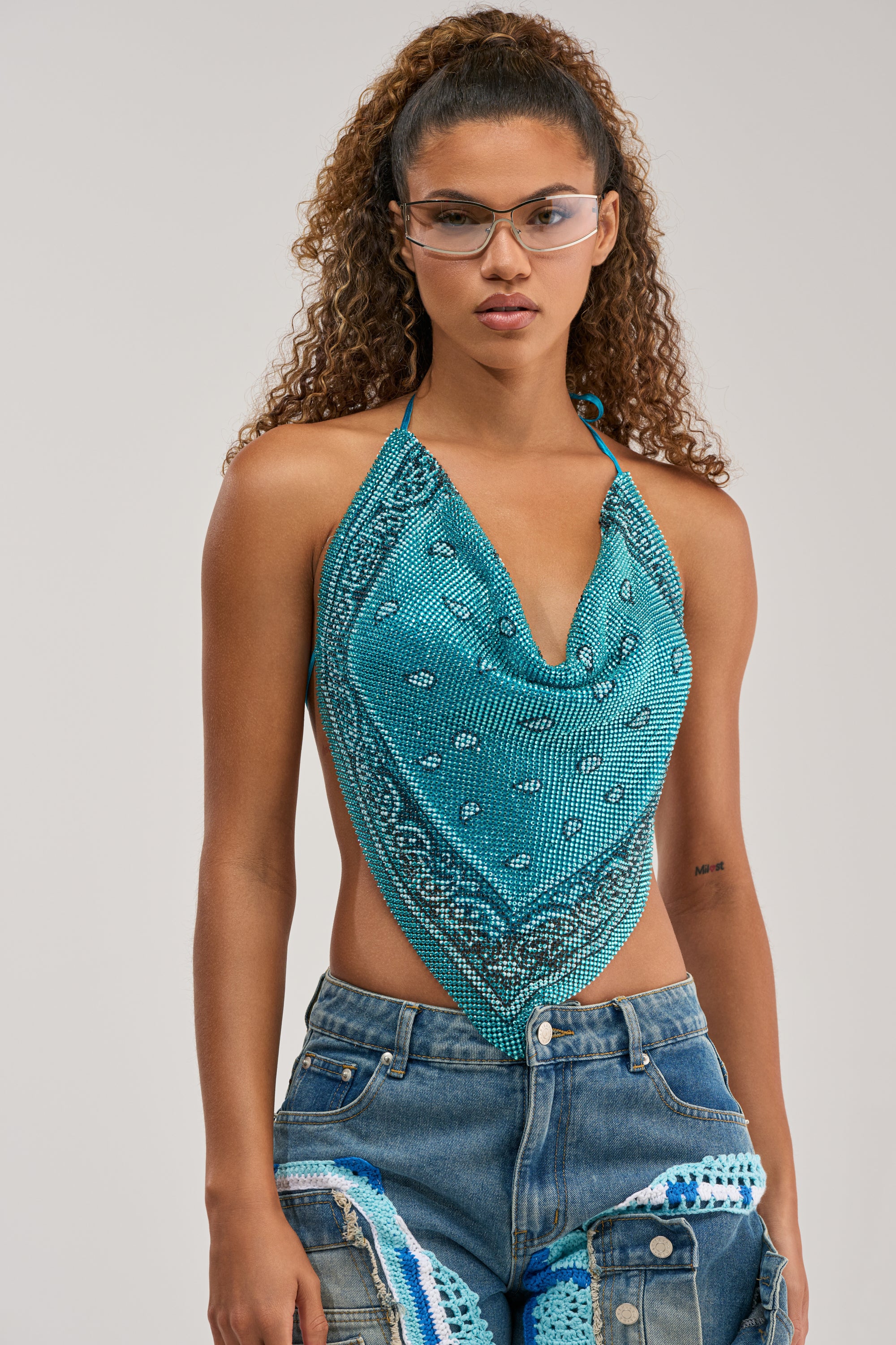 Wearing clear glasses and denim jeans with patchwork details, a woman with curly hair poses confidently against a plain light background. Her concert-ready look is effortlessly chic and modern, highlighted by the FOREVER AND EVER COWL NECK BLOUSE IN LIGHT BLUE styled like a bandana.
