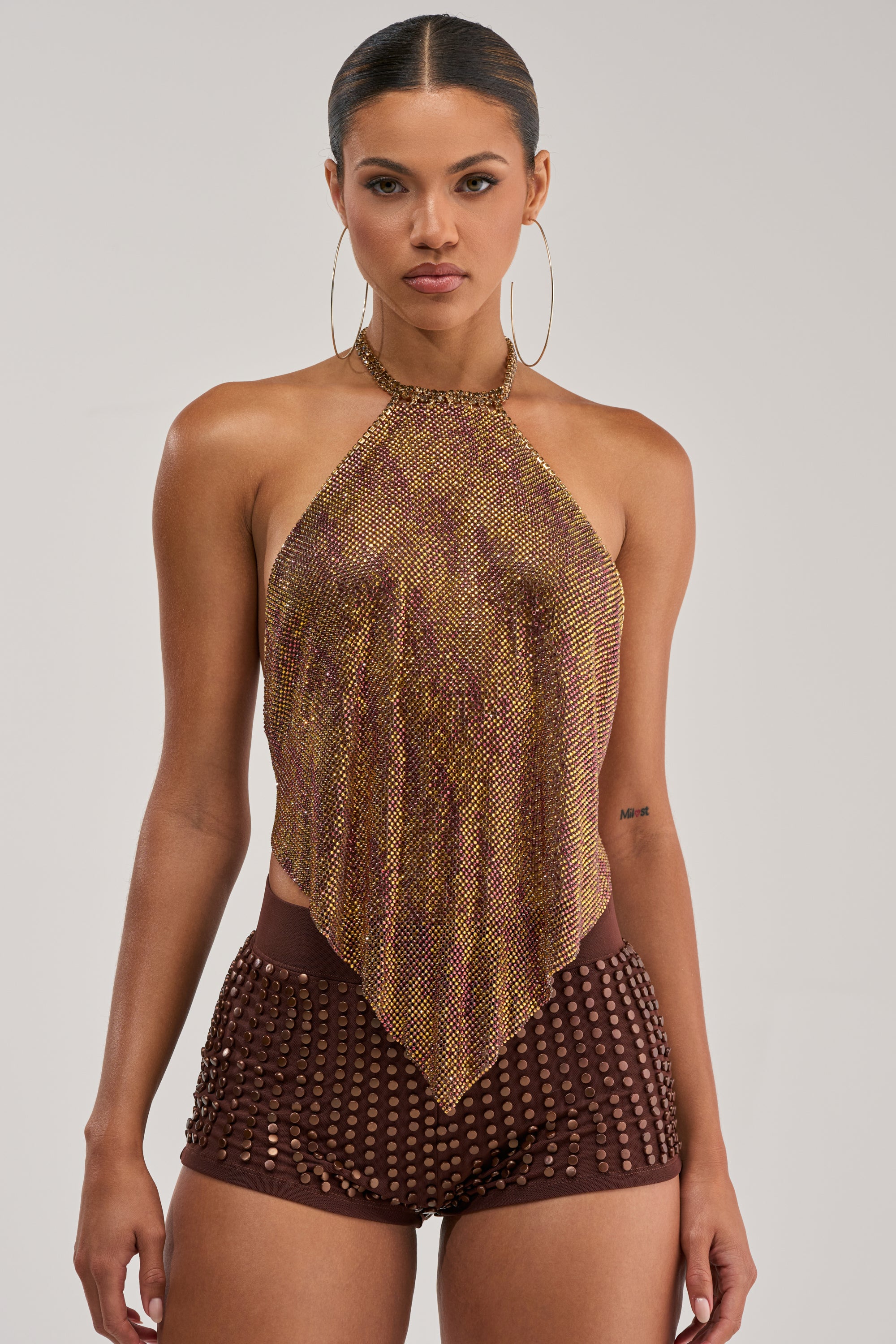 A woman stands facing forward in the GOLDEN GLAM RHINESTONE TOP—a metallic halter with a pointed hem—paired with brown high-waisted shorts with gold studs. She wears slicked-back hair, large hoops, and has a neutral expression.
