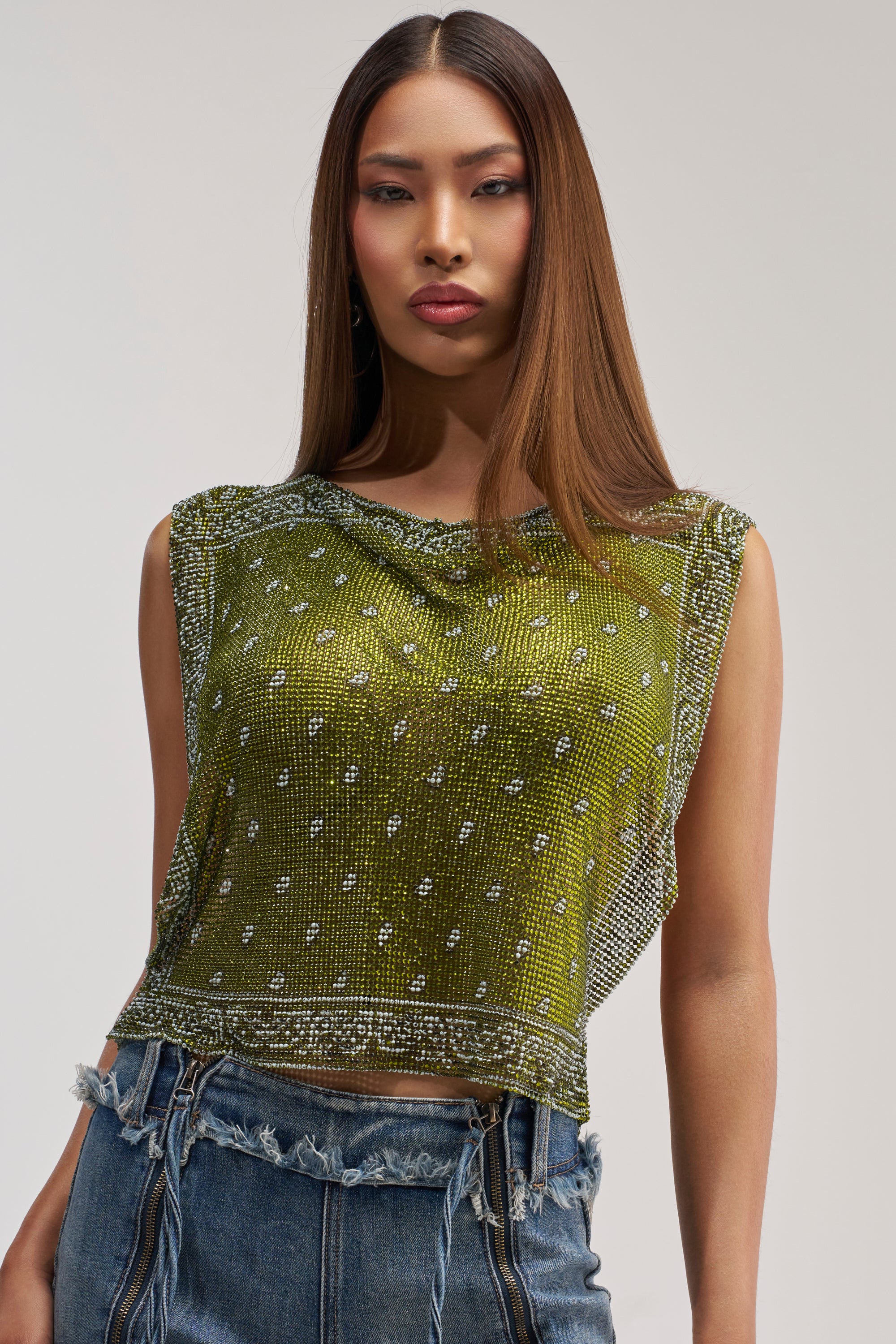 A person wearing the SUMMER LOVIN' RHINESTONE CHAIN TOP—a sleeveless, sheer green festival piece adorned with sparkling rhinestone details—paired with high-waisted denim jeans featuring frayed edges, stands against a plain background.