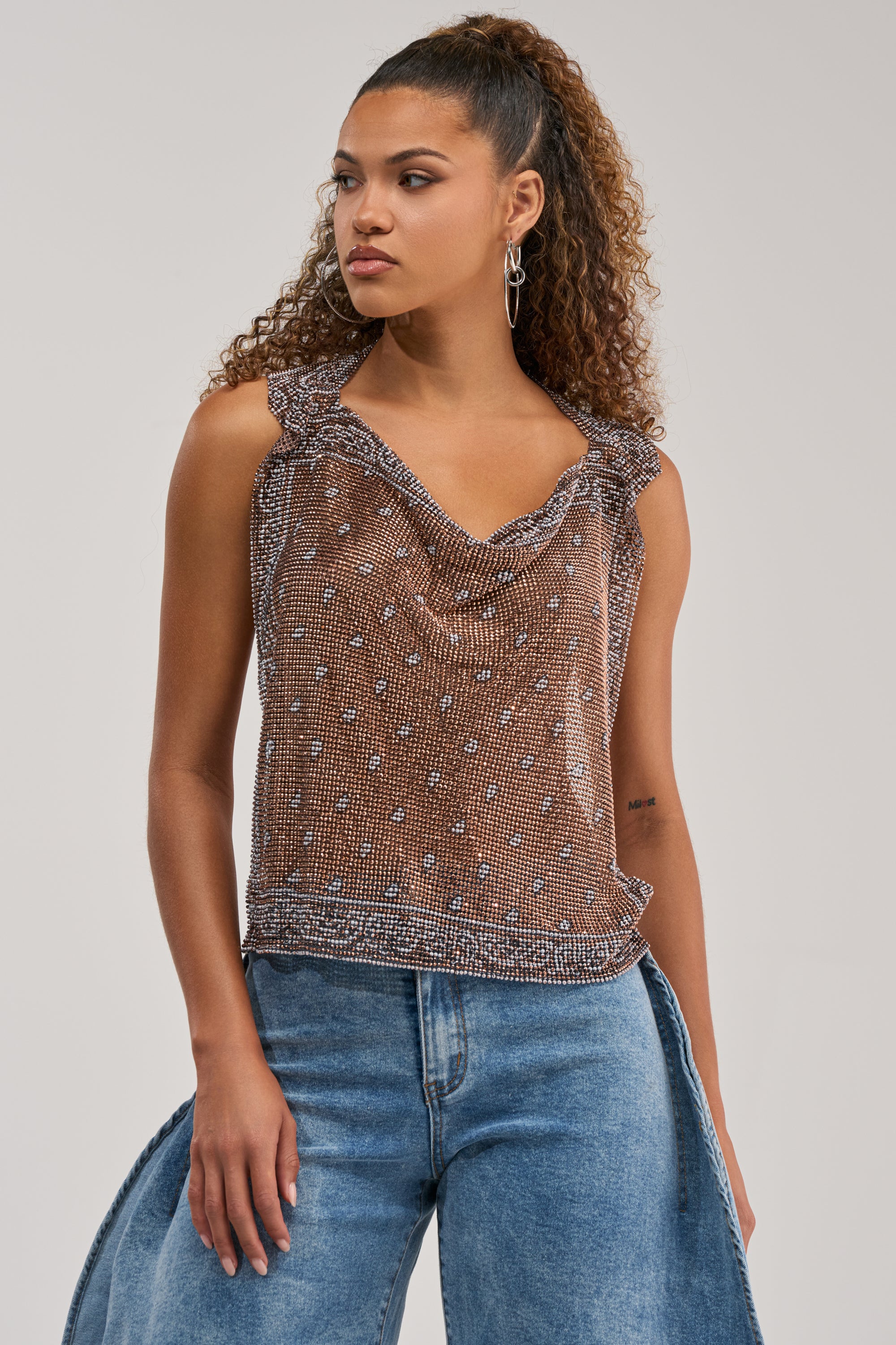 A woman with curly hair in a high ponytail wears the BRONZE BANDANA PRINT RHINESTONE TOP, a brown, sleeveless western festival style with small white dots and ruffle details, paired with blue wide-leg jeans. She faces left with a neutral expression against a plain background.