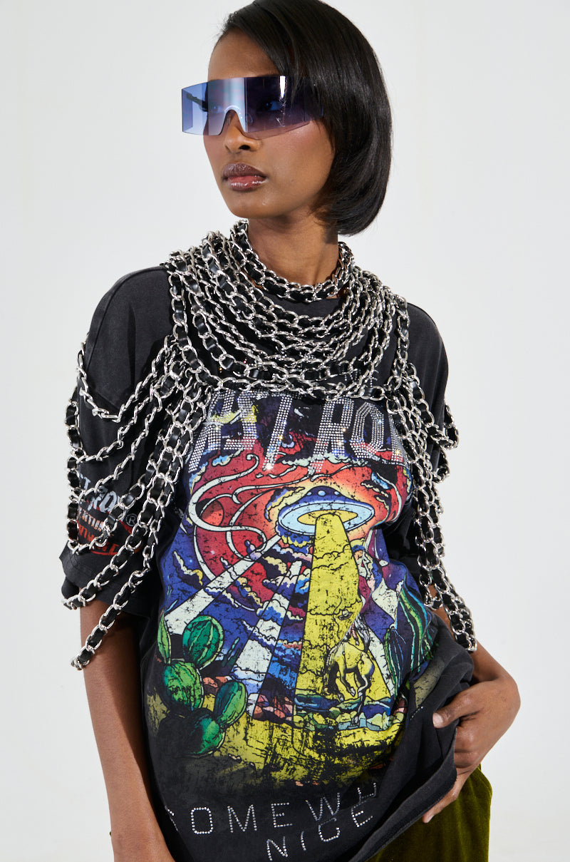 A person wearing a graphic t-shirt with a colorful design, a KYKY Statement Necklace, oversized sunglasses, and dark pants stands against a plain white background. 0